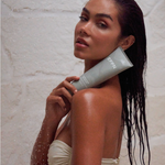 A woman in a beige bikini stands under a shower, holding a gray tube of EFFN body polish. Water droplets are visible in the air against a light stone wall.