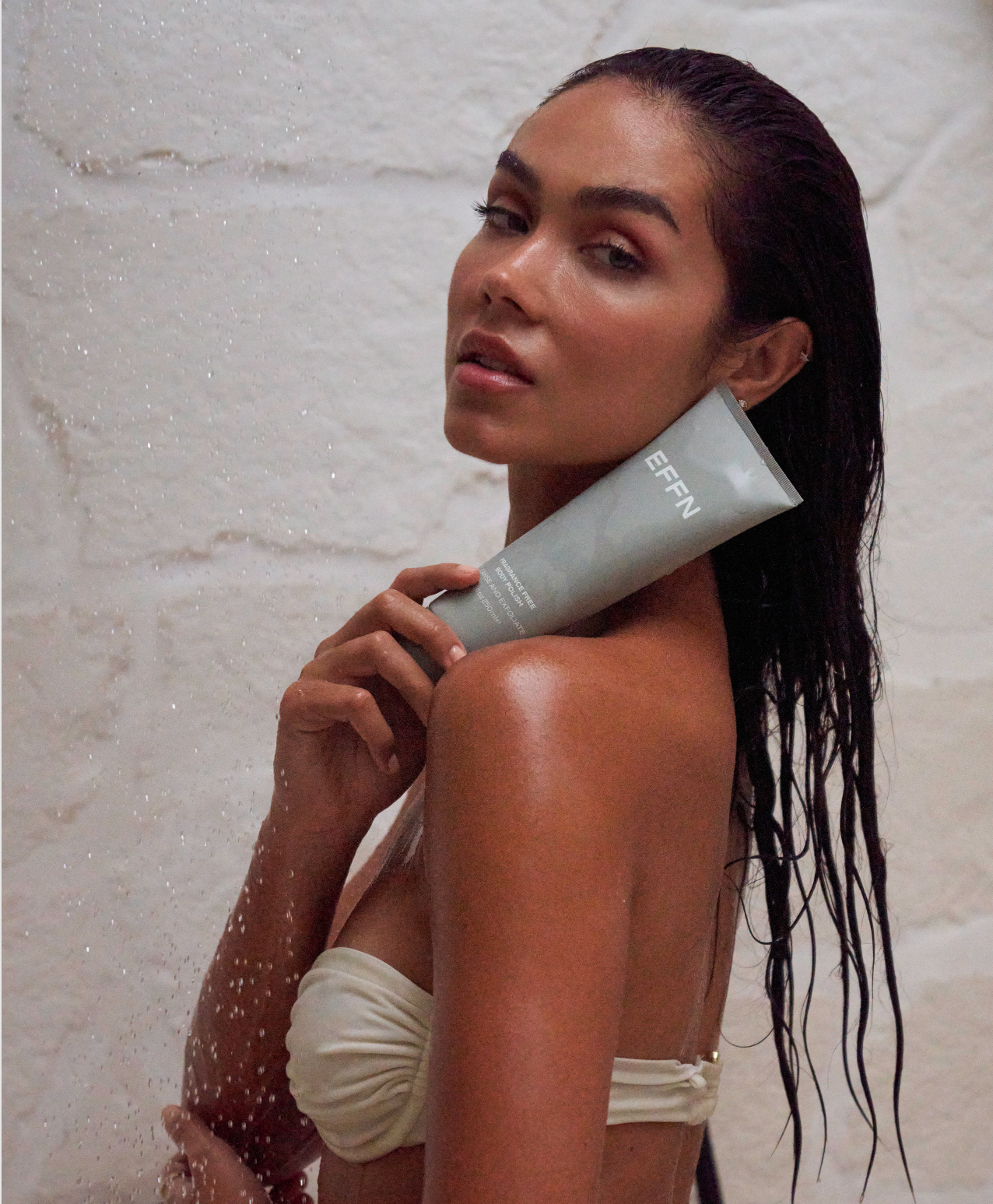 A woman in a beige bikini stands under a shower, holding a gray tube of EFFN body polish. Water droplets are visible in the air against a light stone wall.